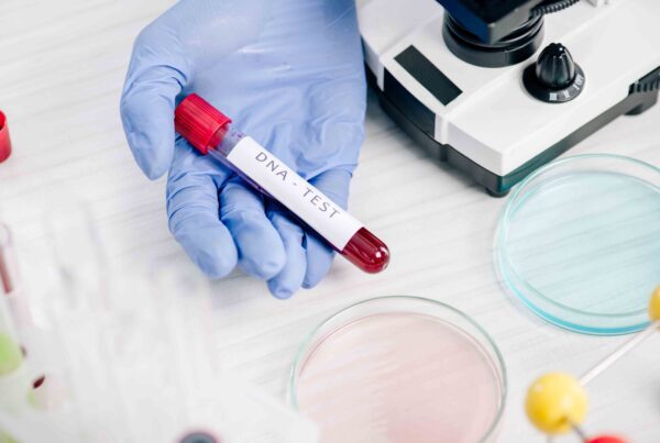 Gloved hand holding DNA test vial next to microscope and petri dishes in a lab.