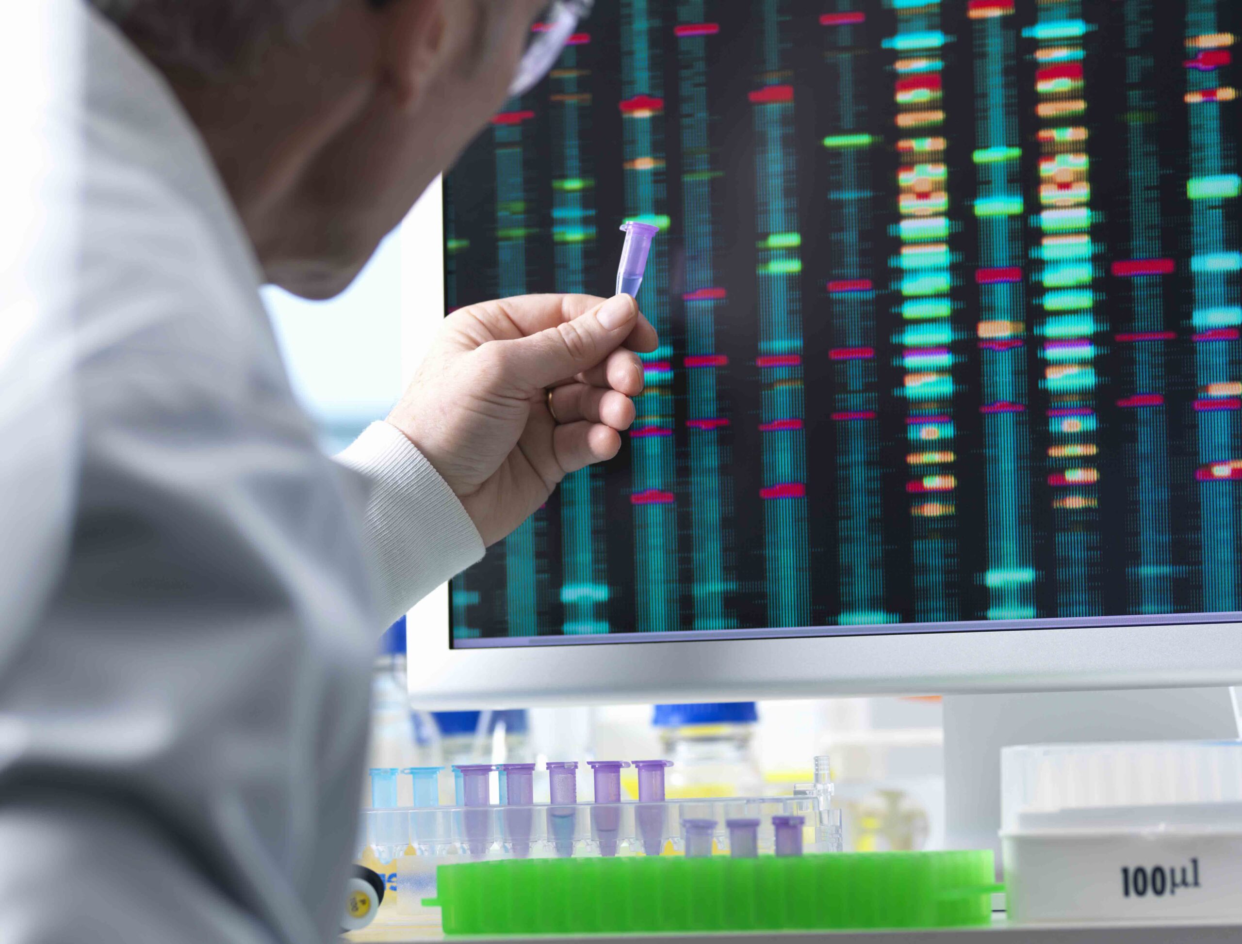 Scientist analyzing DNA sequences on a computer screen, holding a test tube in a lab.