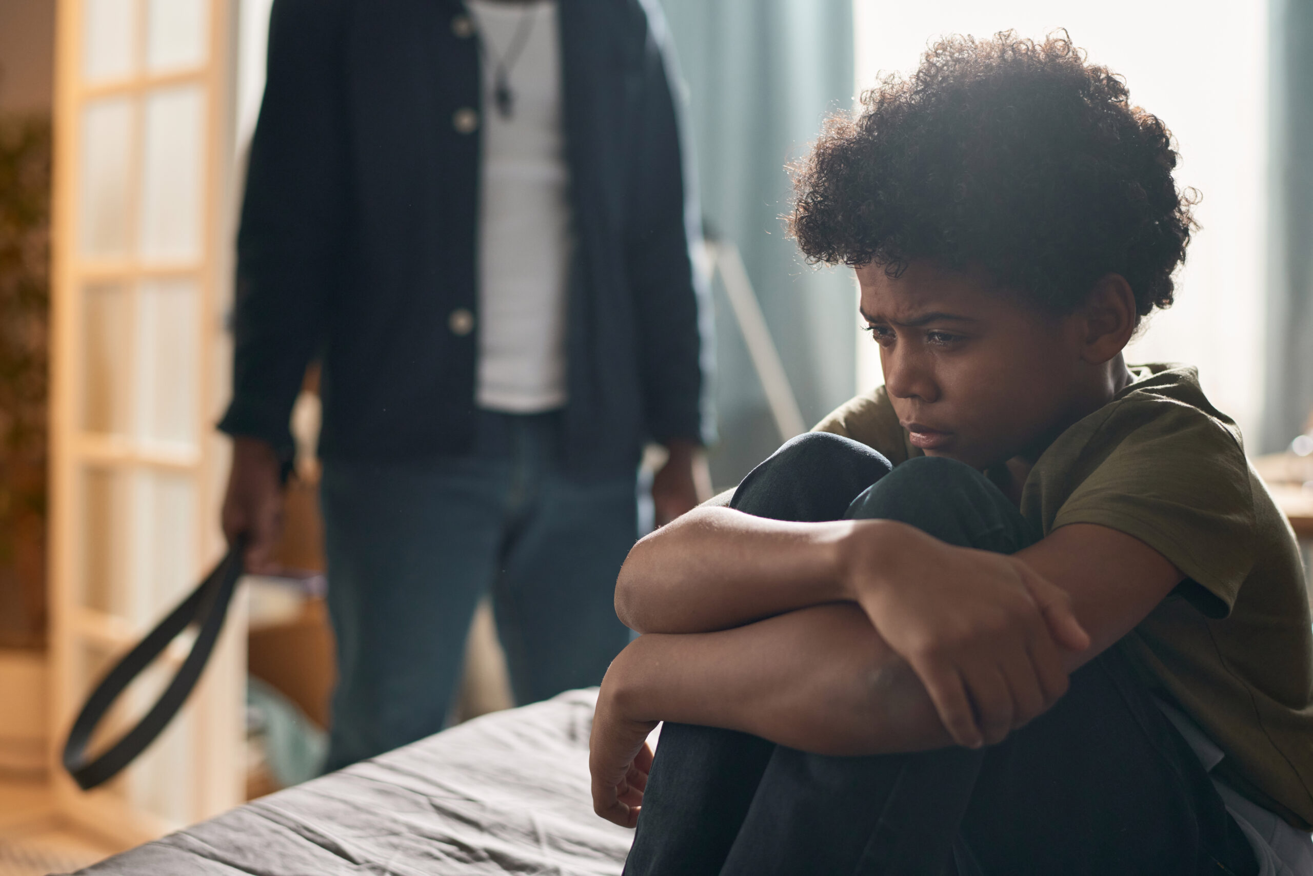 Image of a person sitting on a bed with knees drawn up to the chest, while another person stands nearby holding a belt, highlighting the necessity of legal protection through restraining orders and protective orders