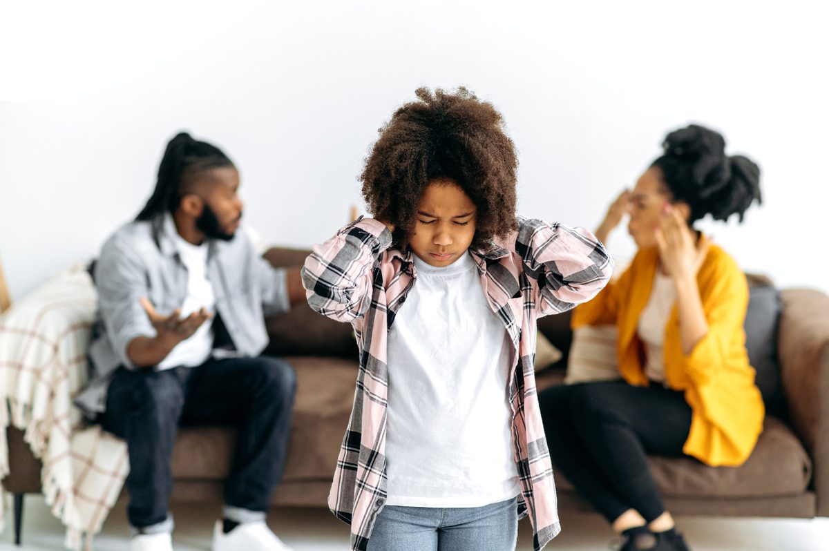 A young child stands in front of a couch, hands on her head, expressing concern or contemplation