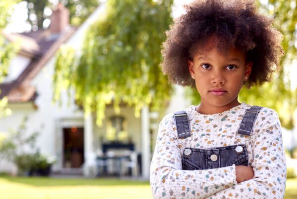 A young girl with an afro stands in front of a house, embodying innocence and joy in a serene neighborhood setting