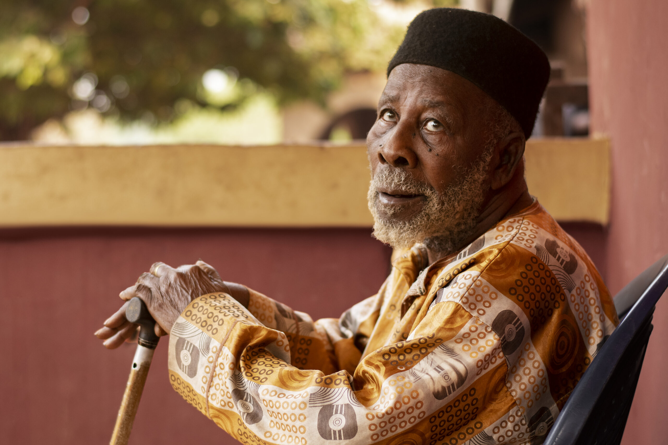 A side-profile portrait of an elderly man with a grey beard and a black cap, sitting outdoors and resting his hands on the handle of a wooden walking cane