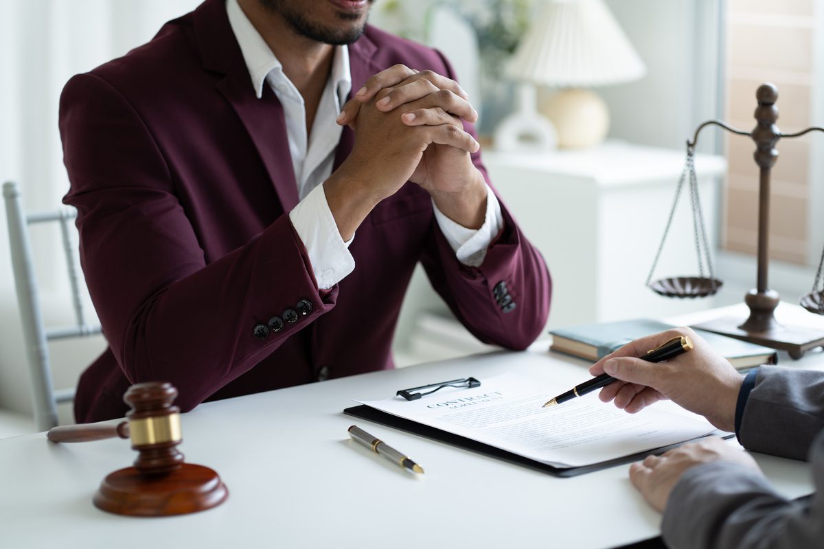 Legal professional in a burgundy suit sitting at a desk with clasped hands while a client reviews a contract agreement