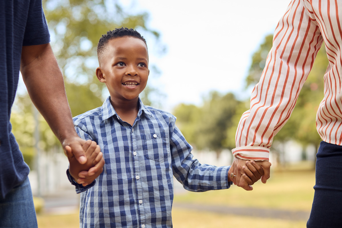 Happy child holding hands with both parents outdoors, representing family unity, child well-being, parental support, co-parenting, and strong family relationships.