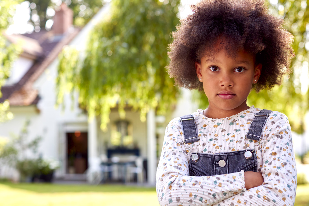 Young child standing in front of a family home with arms crossed, symbolizing child adoption, foster care, child welfare, family law services, and providing a safe and permanent home for children.